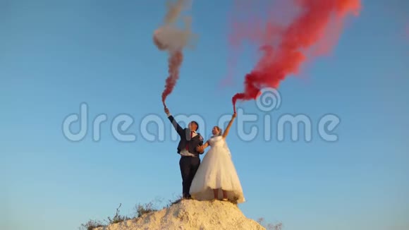 幸福的新娘和新郎在蓝天上挥舞着粉红色的烟笑着蜜月浪漫两者之间的关系视频的预览图