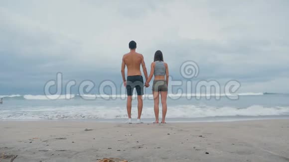 年轻的一家人牵着手站在海滩的海洋前旅途中享受海浪的年轻夫妇视频的预览图