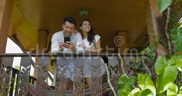 年轻夫妇在阳台上抬头望着说话的男人和女人拥抱着用手机开心地笑着视频的预览图