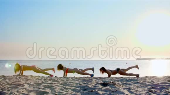 三个漂亮运动的女人穿泳衣做俯卧撑同步练习在海滩上夏天在视频的预览图