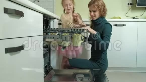 可爱的金发女婴穿着粉红色的裙子在明亮的厨房里帮助妈妈从洗碗机里拿出盘子和杯子视频的预览图