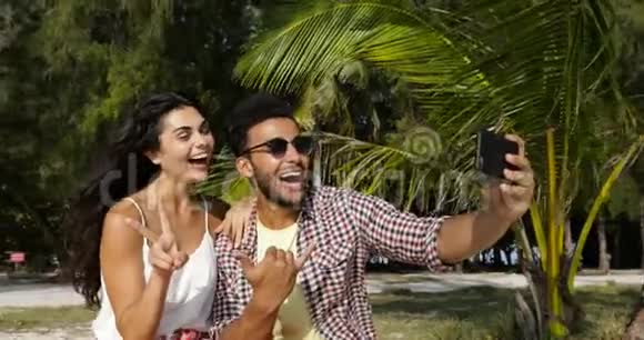 情侣使用手机在海滩棕榈树下自助拍照快乐的微笑男人和女人视频的预览图
