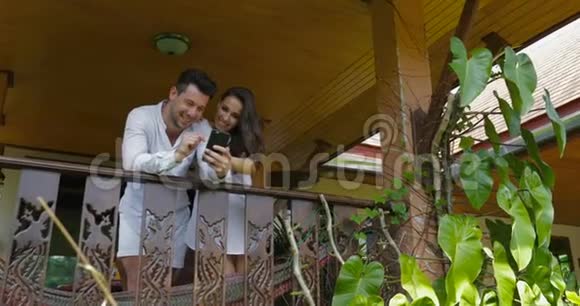 年轻夫妇在酒店的阳台上拿着手机快乐的男人和女人一起上网视频的预览图