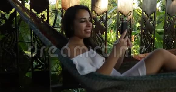 年轻女子触摸手机智能手机躺在吊床上在夏日露台上传递信息快乐微笑女孩视频的预览图