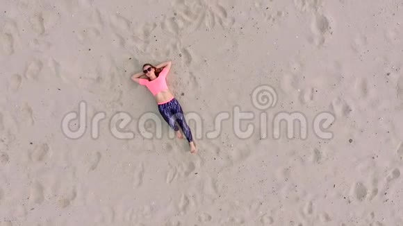时尚的年轻运动美女戴着太阳镜躺在沙滩上在一个荒芜的海滩上夏天从上面看视频的预览图