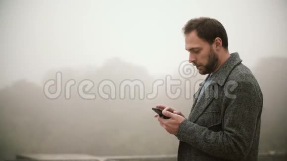 年轻英俊的男子站在公园雾蒙蒙的早晨长胡子的男性使用带有触摸屏的智能手机视频的预览图