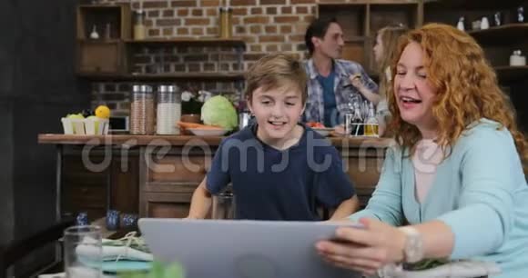 母亲和儿子一起举起手提电脑制作视频电话让父亲和女儿在厨房做饭视频的预览图