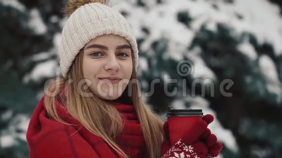 年轻漂亮的女孩穿着温暖的衣服站在圣诞树附近在雪地里喝着报纸上的热饮料视频的预览图