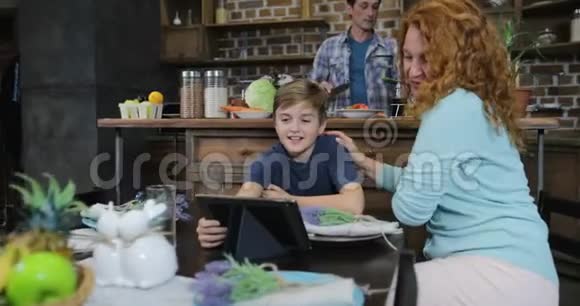 母亲和儿子在父亲和女儿一起享用蔬菜晚餐时使用数字平板厨房沙拉视频的预览图