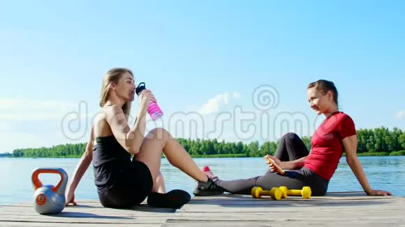 两个漂亮的运动的金发女孩女人在户外训练后休息喝水说话笑湖泊河流视频的预览图