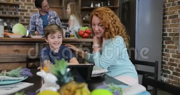 母亲和儿子触摸平板电脑屏幕浏览食谱而父亲与女儿切蔬菜晚餐沙拉视频的预览图
