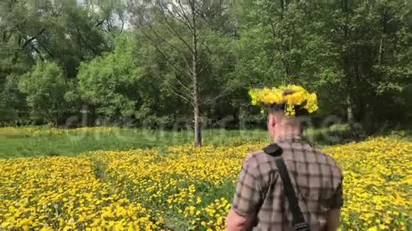 一个年轻人走在春天的草地上长满了开花的蒲公英头上是蒲公英的花环阳光明媚的春天视频的预览图