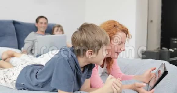 妈妈和儿子一起笑看数码平板电脑上的搞笑视频躺在床上快乐的一家人在一起视频的预览图