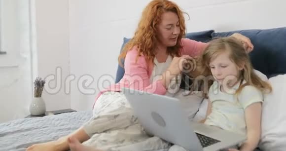 母亲在女孩使用笔记本电脑时抚摸女儿的头一家人坐在卧室的床上在一起视频的预览图