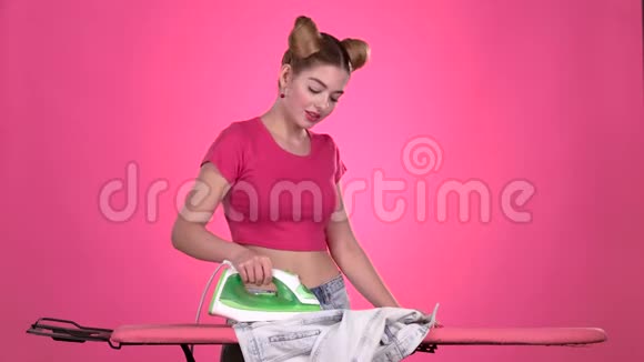 女孩在粉红色的板上用熨斗熨烫衣服粉色背景视频的预览图