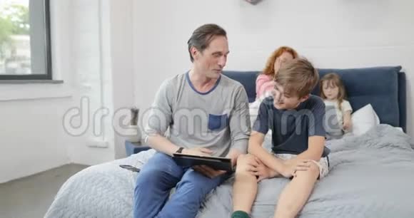 父亲和儿子用数码平板电脑坐在母亲的床上女儿抱着笔记本电脑家庭在一起视频的预览图