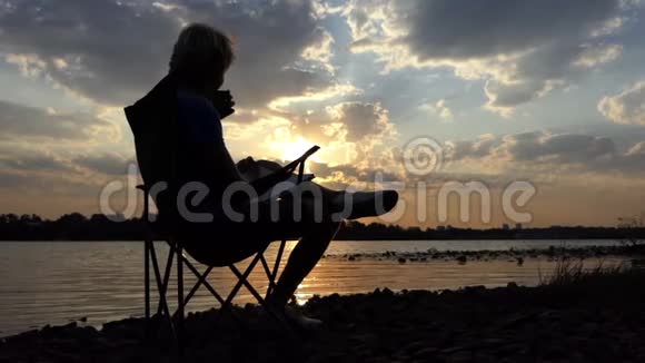男人坐在河岸边的椅子上在斯隆莫的日落时看书和喝酒视频的预览图