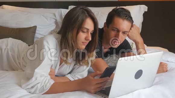 年轻夫妇穿着浴袍躺在床上手提电脑视频的预览图