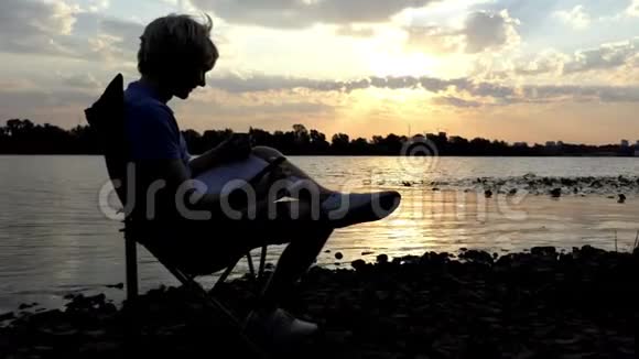 年轻人坐在夕阳下的河岸边的椅子上写着微笑视频的预览图
