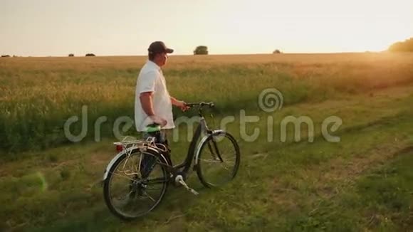农夫在麦田的背景下骑着自行车沿着乡间的道路行驶傍晚日落时分慢吞吞视频的预览图