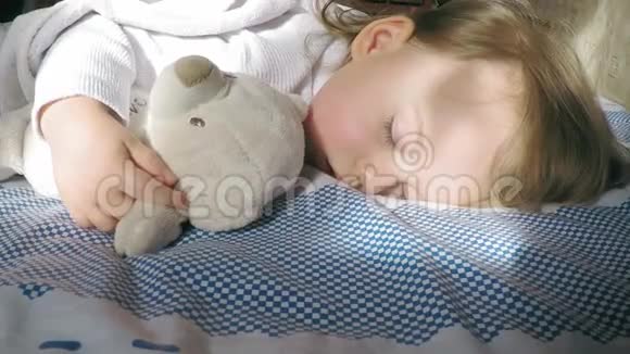 可爱的小女孩金发睡在床上被太阳照亮带着泰迪熊视频的预览图