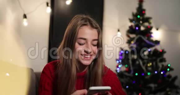 新年庆典穿着红色毛衣的迷人年轻女子坐在圣诞树前的沙发上和她聊天视频的预览图