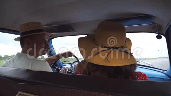 年轻夫妇戴着帽子在夏季旅行时坐着老式汽车男人和女人坐在旧复古的前座上视频的预览图
