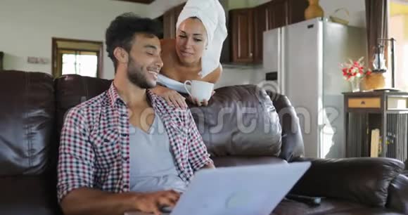 女人对坐在教练上的男人说话使用笔记本电脑在客厅在线浏览年轻夫妇在早晨视频的预览图