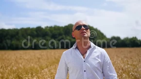 一个穿着白色衬衫的秃头站在金色麦田里的肖像在阳光明媚的天空背景上歌唱视频的预览图