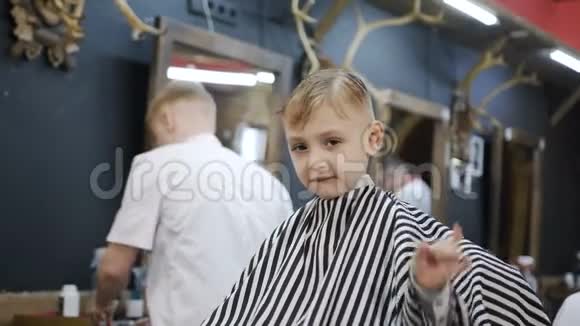 一个小男孩坐在理发店的椅子上看着镜头用手显示一切都很好他视频的预览图
