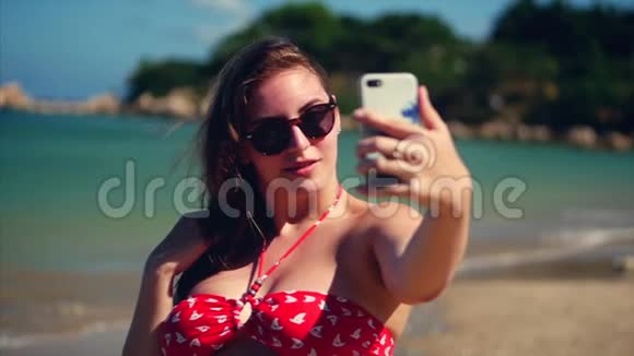 年轻美丽的布鲁内特女士站在沙滩上夏天看着她的手机和自拍背景蓝色视频的预览图