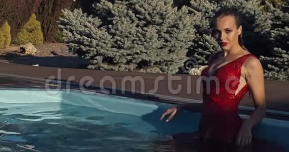 一位身穿红色连衣裙化妆和湿发的年轻女子正在游泳池里摆姿势4K视频的预览图