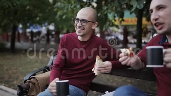 坐在公园长椅上的男人吃三明治和薯条喝杯子里的咖啡和聊天视频的预览图