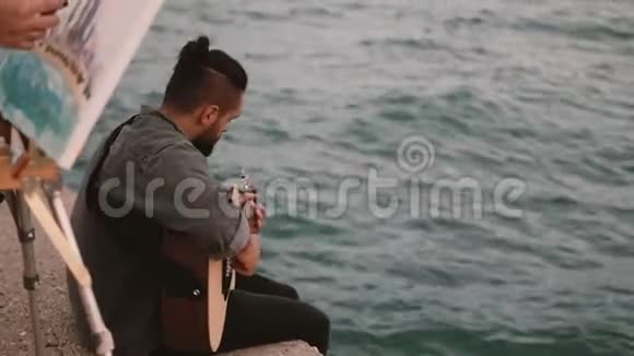 年轻的天才夫妇坐在海边做着业余爱好女人画画男人弹吉他视频的预览图