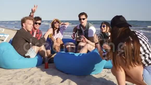 年轻的时髦女孩录制视频或拍摄一群朋友坐在沙滩上的安乐椅上玩耍视频的预览图