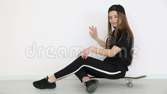 少女穿着时髦的嘻哈服装戴着帽子用滑板摆在白色的墙上视频的预览图