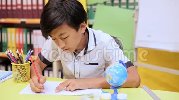 一个男孩坐在桌子旁用红色铅笔在笔记本上写字视频的预览图