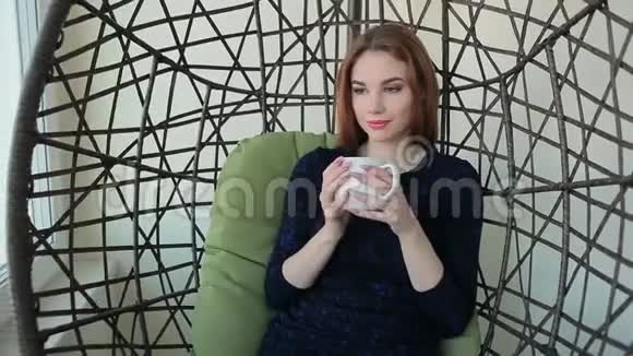 美丽的年轻女子坐在挂椅上端着一杯热茶或咖啡视频的预览图