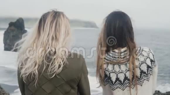 两个旅行的女人站在黑色的海滩上在冰岛熊怪的脚趾岩石看着水微笑玩得开心视频的预览图