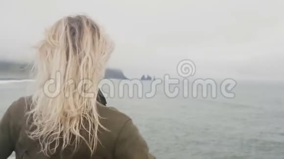 冰岛黑滩上站着一位年轻的金发女子的后景自由游客举起双手随风飘扬视频的预览图