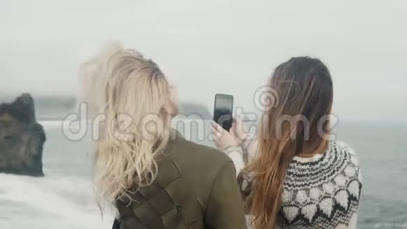 两个快乐的女人在智能手机上自拍照片女孩们花时间在冰岛巨魔脚趾附近的海滩上视频的预览图