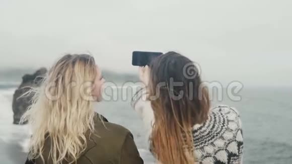 后景两位年轻女性在冰岛巨魔脚趾岩石附近的海边自拍照片视频的预览图