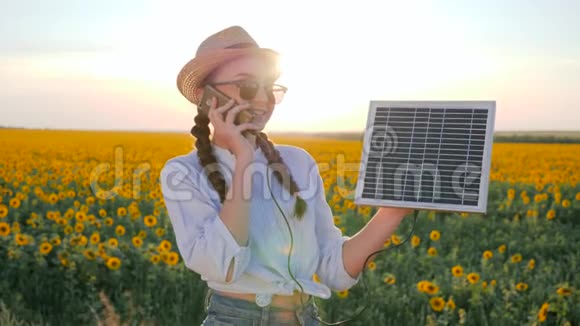 女孩用手机说话拿着太阳能电池板在向日葵的背景地里年轻女子拿着手机和太阳能视频的预览图