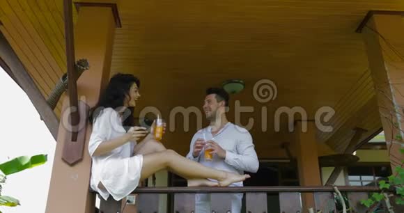 年轻夫妇在酒店阳台上聊天快乐的男女拿着橙汁杯子交流视频的预览图