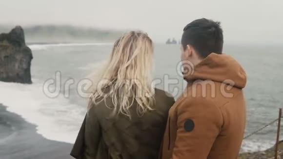 年轻的旅行夫妇在冰岛的黑色海滩上看着巨怪的脚趾岩石著名的景象感觉快乐举手视频的预览图