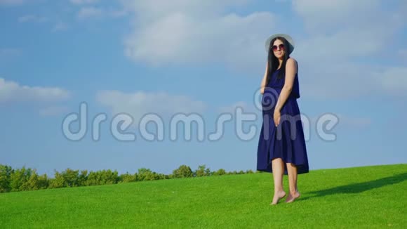 一个戴着帽子和太阳镜等孩子的时髦女人站在蓝天背景下的绿色草地上视频的预览图