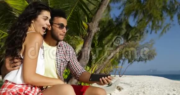情侣坐在沙滩上用手机拥抱快乐的微笑男人和女人交谈视频的预览图