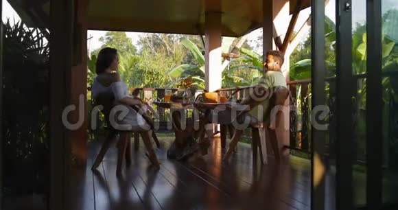 年轻夫妇坐在露台上享受日落的热带森林景观谈论和碰撞果汁眼镜快乐的人和视频的预览图