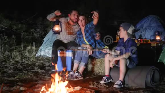 营地幸福的家庭在森林篝火附近的手机上录制视频爸爸妈妈带着煤油灯视频的预览图