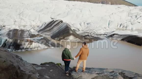 年轻美丽的一对夫妇站在冰岛的瓦特纳霍卡尔冰湖的岸边看着冰川视频的预览图